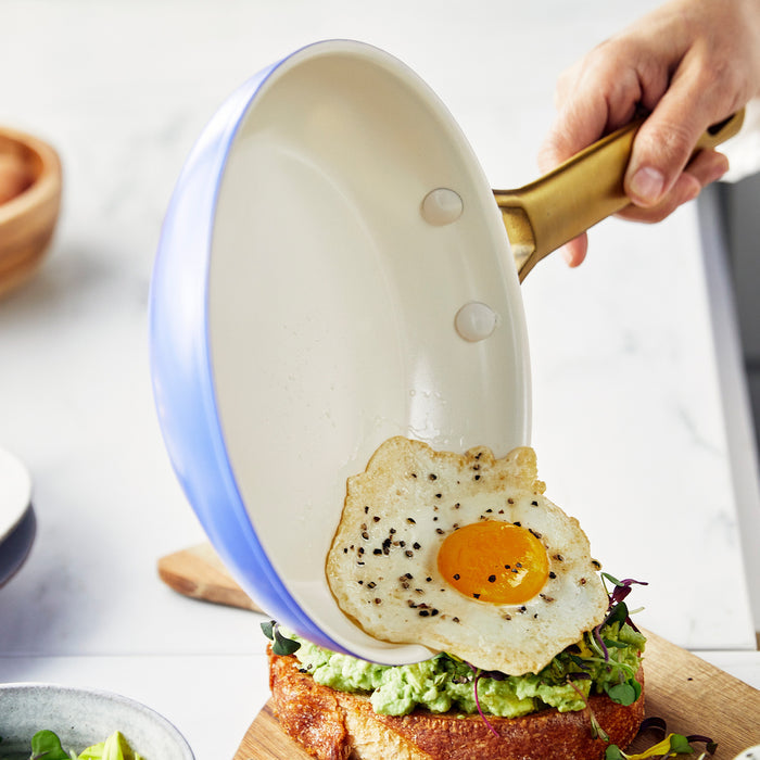 Hand sliding a sunny-side-up egg from a purple ceramic nonstick frypan onto avocado toast.