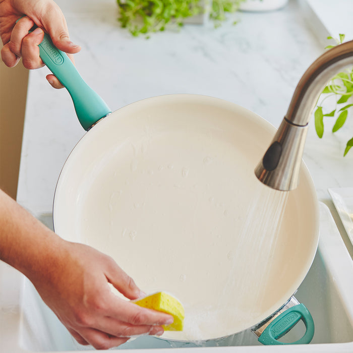 Turquoise ceramic frypan being washed under running water with a yellow sponge.