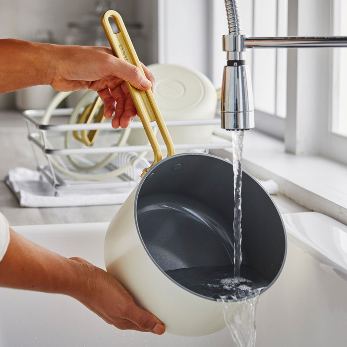 White ceramic saucepan with lid being washed at a sink, other dishes drying in the background.