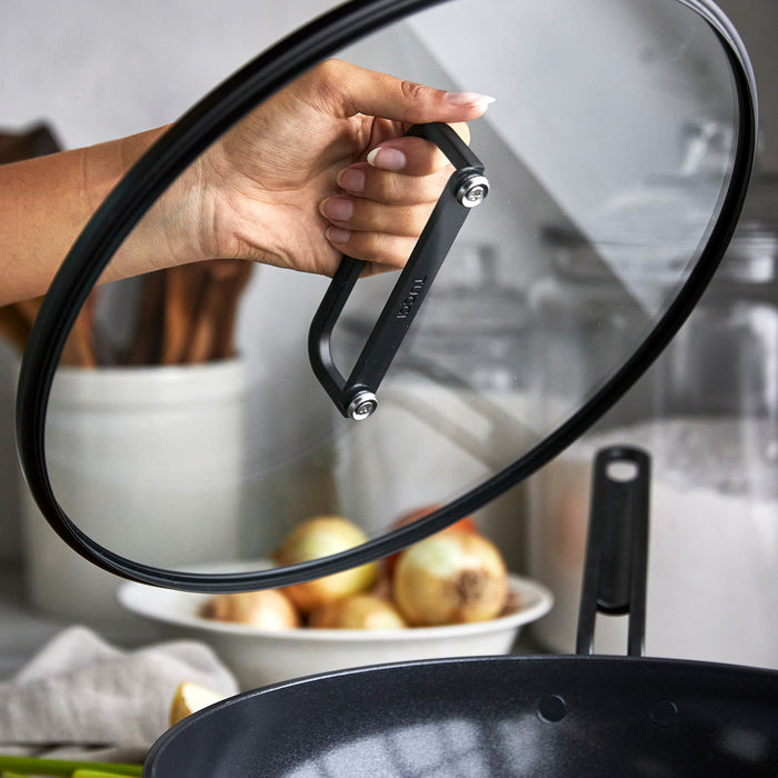 Hand lifting glass lid from black ceramic nonstick 6.5-quart pan on a kitchen counter.