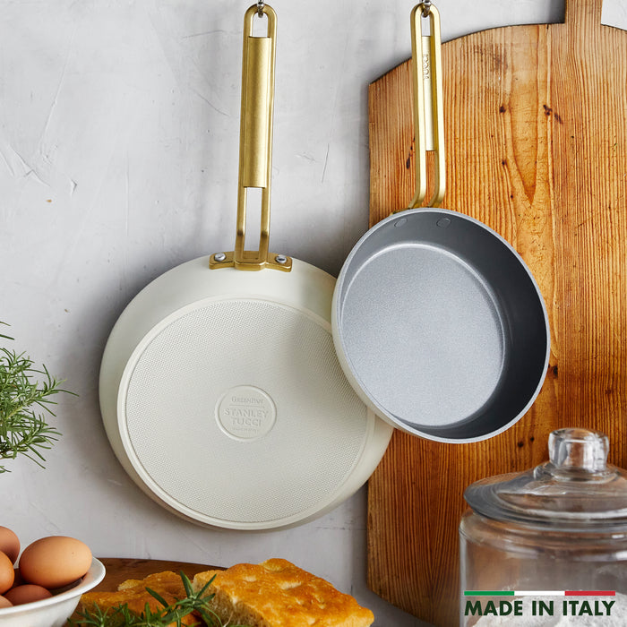 Two ceramic nonstick frypans hang above a table with bread, eggs, herbs, and a Made in Italy sign.