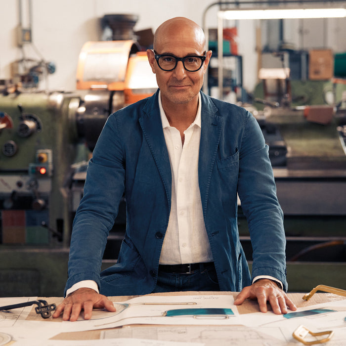 Man in glasses and blue jacket stands behind a table displaying Stanley Tucci saucepan design sketches.