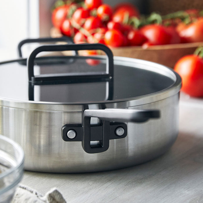 Stainless steel ceramic sauté pan with lid on counter, tomatoes visible in the background.