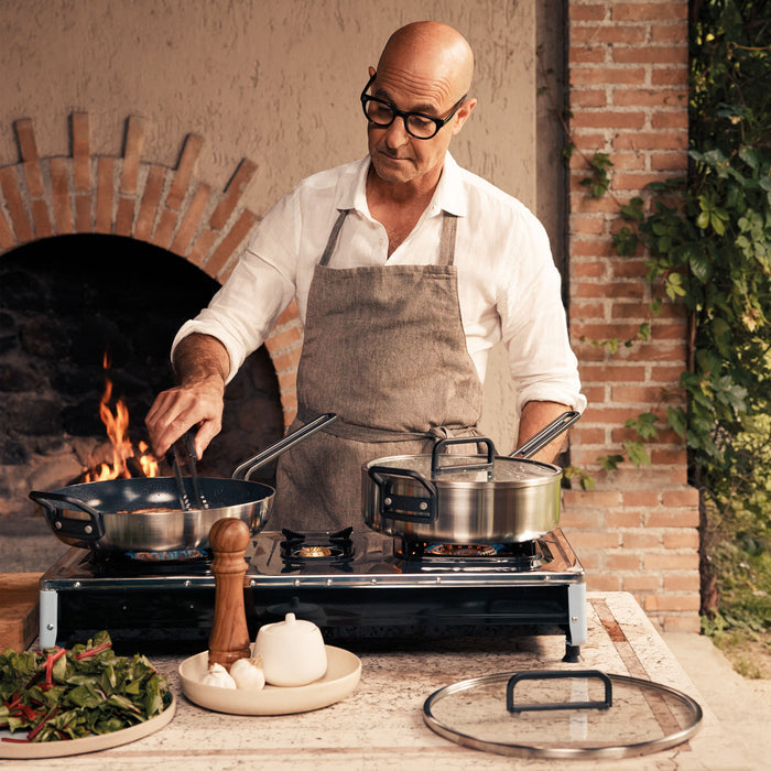 Man cooking outdoors with a stainless steel sauté pan and fresh ingredients, wearing an apron.