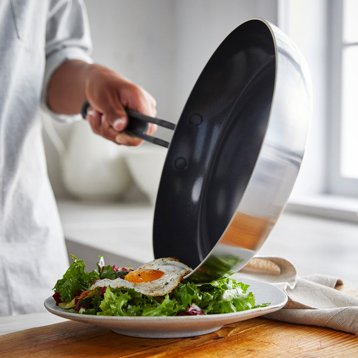 Fried egg being slid from a ceramic nonstick frypan onto a plate of leafy salad on a kitchen counter.
