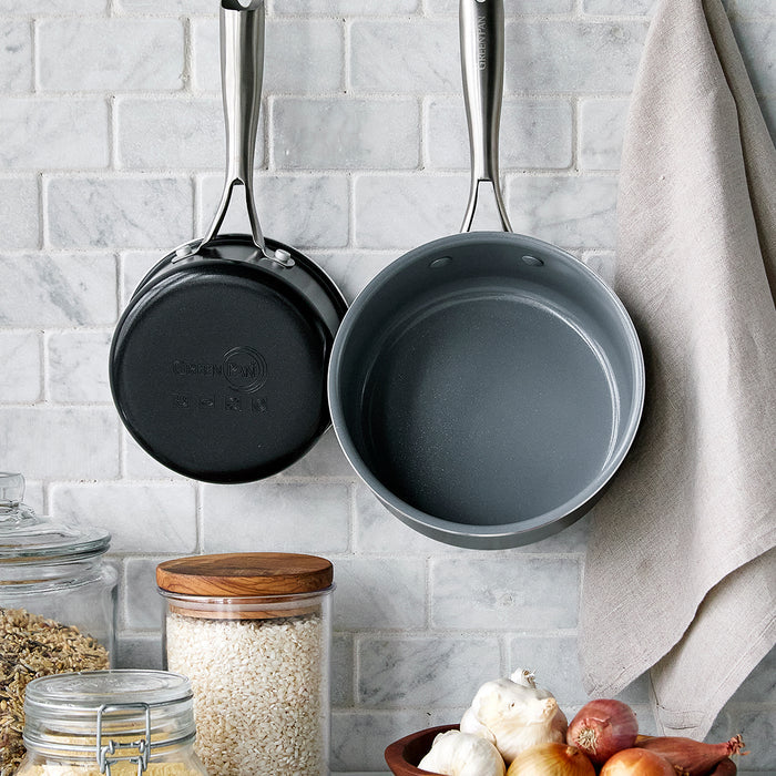 Two ceramic nonstick saucepans, 1-quart and 2-quart, hang on a tile wall above jars of grains, garlic, and onions.