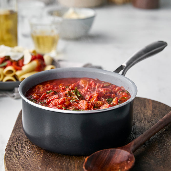 Ceramic nonstick saucepan set on a wooden board, pasta dish blurred in the background.