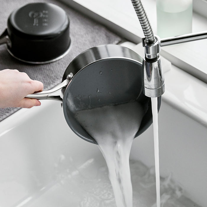 Hand pouring water from a ceramic nonstick saucepan into a sink under a running tap.