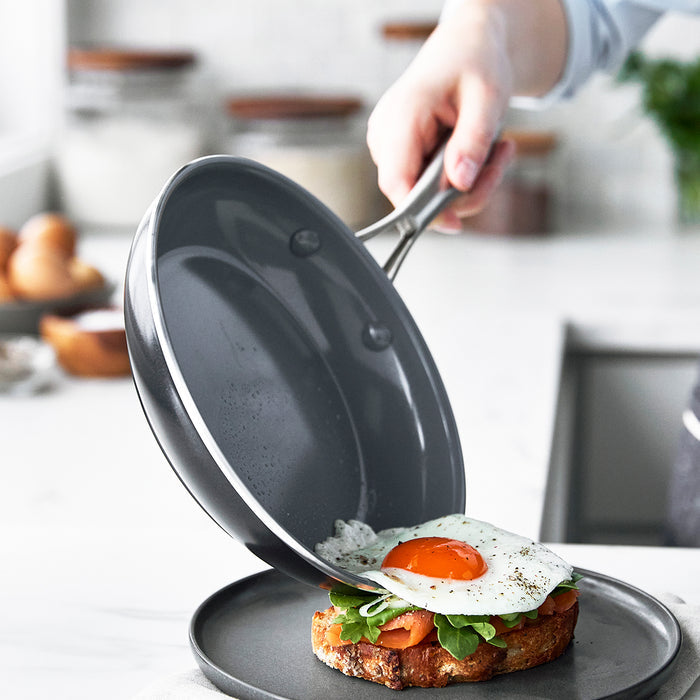 A hand slides a fried egg from a ceramic nonstick frypan onto toast with greens and tomatoes.