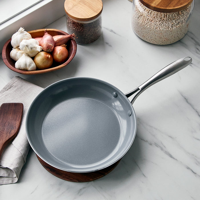 Ceramic nonstick 10-inch frypan on marble countertop with garlic, onions, jars, and wooden spatula nearby.