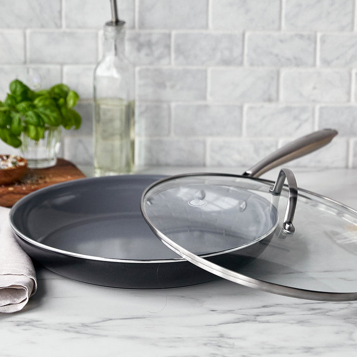 Ceramic nonstick 12-inch frypan with glass lid on marble counter, next to basil, a loaf of bread, and a bottle.