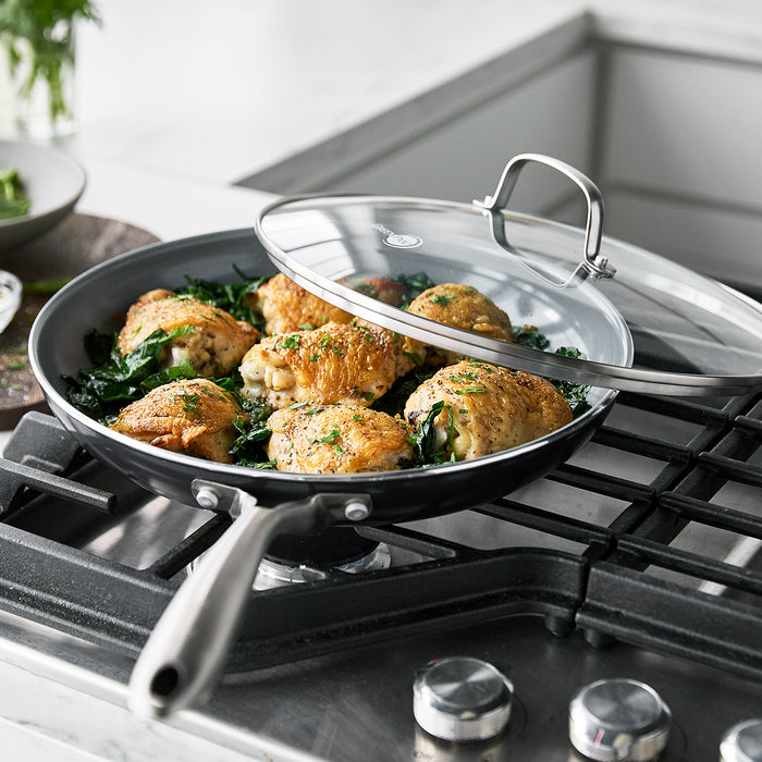Chicken thighs and greens cooking in a 12-inch ceramic nonstick frypan with a partially covered glass lid.