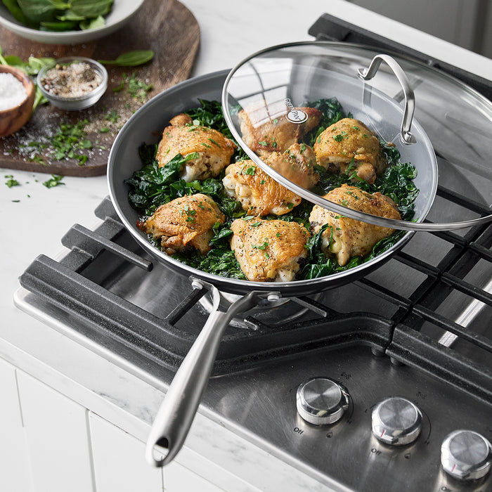Four cooked chicken thighs on greens in a ceramic nonstick 12-inch frypan with lid on a gas stove.