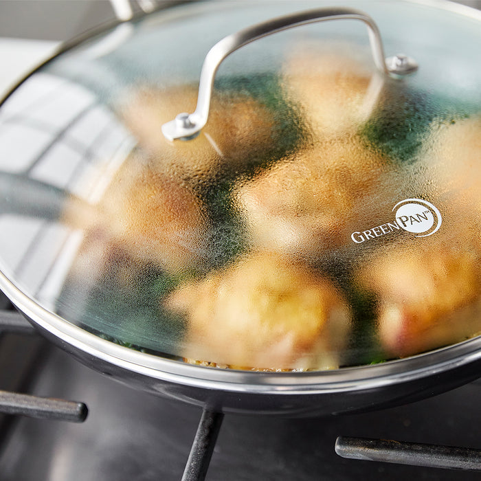 Swift ceramic nonstick 12-inch frypan with glass lid cooking chicken, GreenPan logo on stainless steel handle.