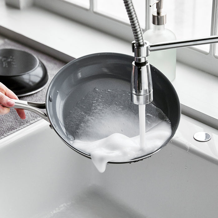 Hand washing a ceramic nonstick frypan with lid under running water in a kitchen sink by a window.