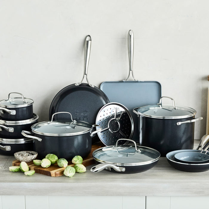 Black ceramic nonstick cookware set with metal handles and glass lids, 17 pieces, some filled with Brussels sprouts.