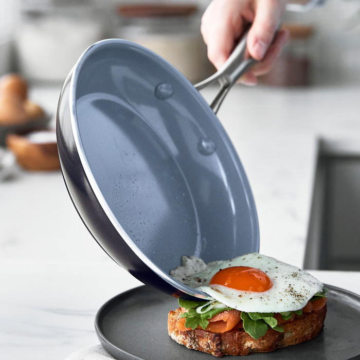 A hand slides a fried egg from a gray ceramic nonstick skillet onto toast and greens on a plate.