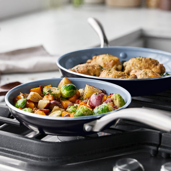Two ceramic nonstick pans from a 17-piece cookware set on a stove, cooking vegetables and chicken thighs.