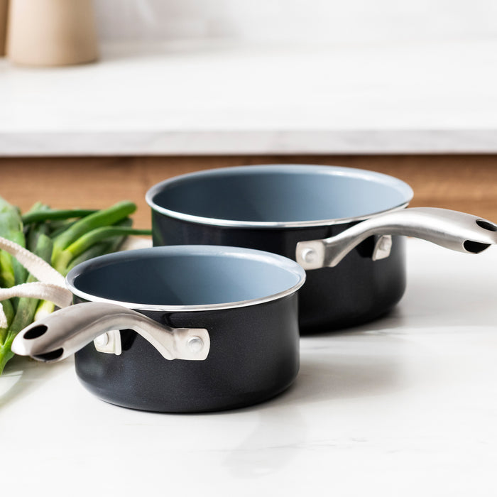 Set of two black ceramic nonstick saucepans with silver handles on a white counter, green onions in background.