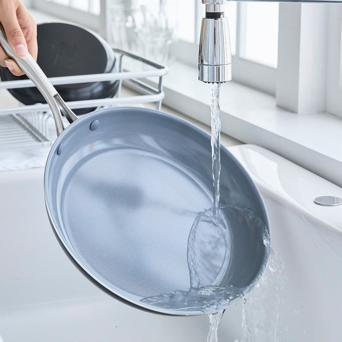 Hand rinsing a ceramic nonstick pan from the 17-piece set under a kitchen faucet in a bright modern kitchen.