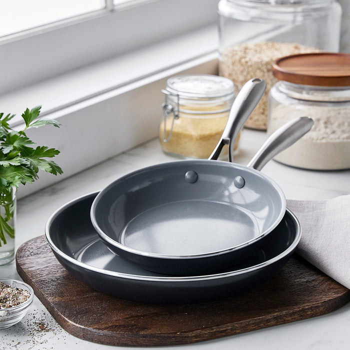 Two gray ceramic nonstick frypans, 8 and 10, stacked on a wooden board with jars and fresh herbs in the background.