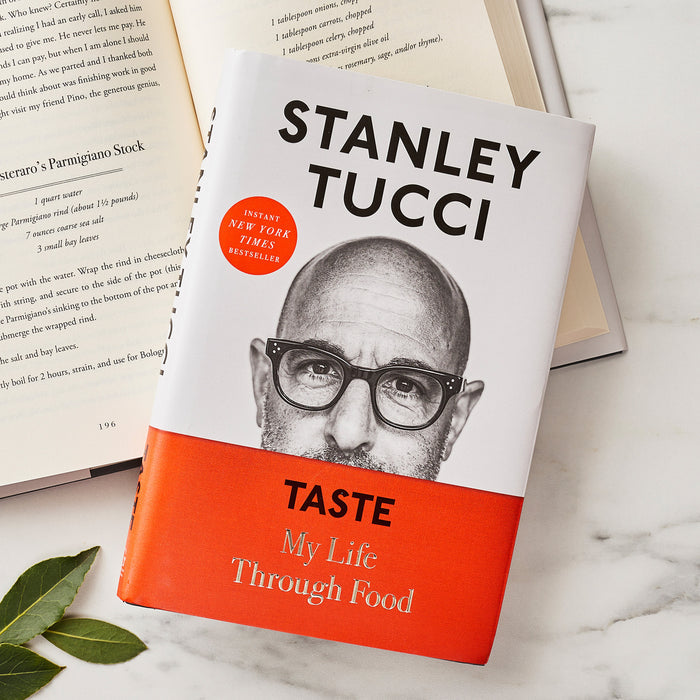 Stanley Tucci’s Taste: My Life Through Food cookbook open on a white surface with green leaves nearby.