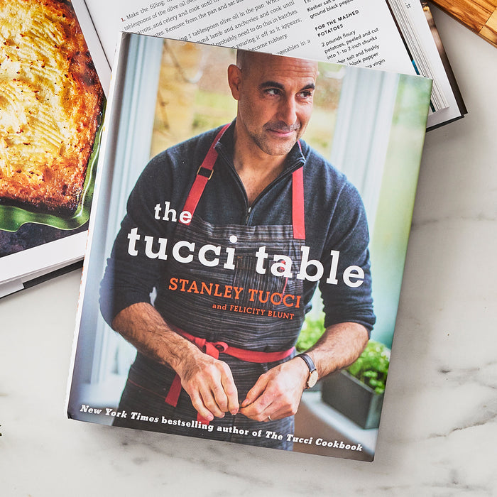 Stanley Tucci stands in a striped apron holding a knife, featured on The Tucci Table Cookbook cover.