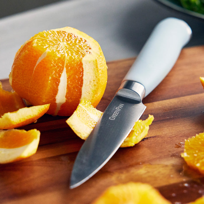 Chef’s knife peeling an orange on a wooden cutting board with orange slices and peels surrounding it.