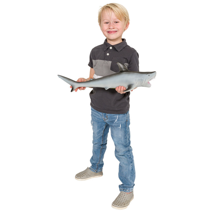 A young boy with blonde hair, in a dark polo shirt and jeans, smiles while holding the Toysmith Epic Shark - Hammerhead, a soft plastic shark toy by Toysmith, against a white background.