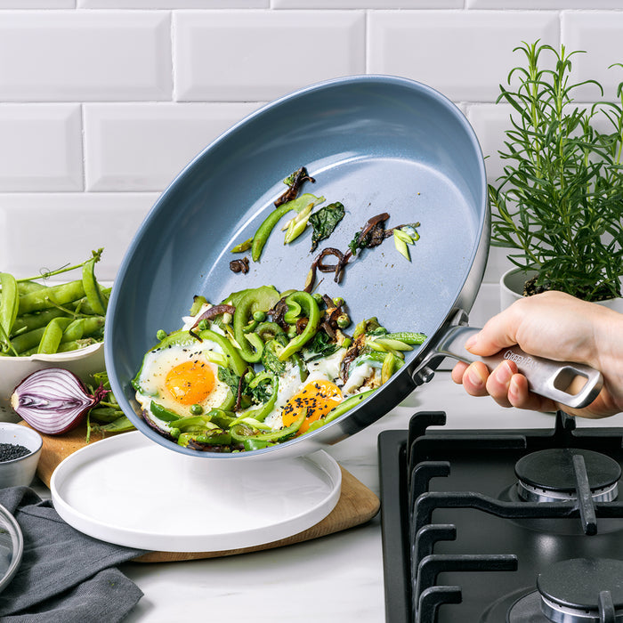 Hand holding a ceramic skillet with eggs and vegetables by the stovetop.