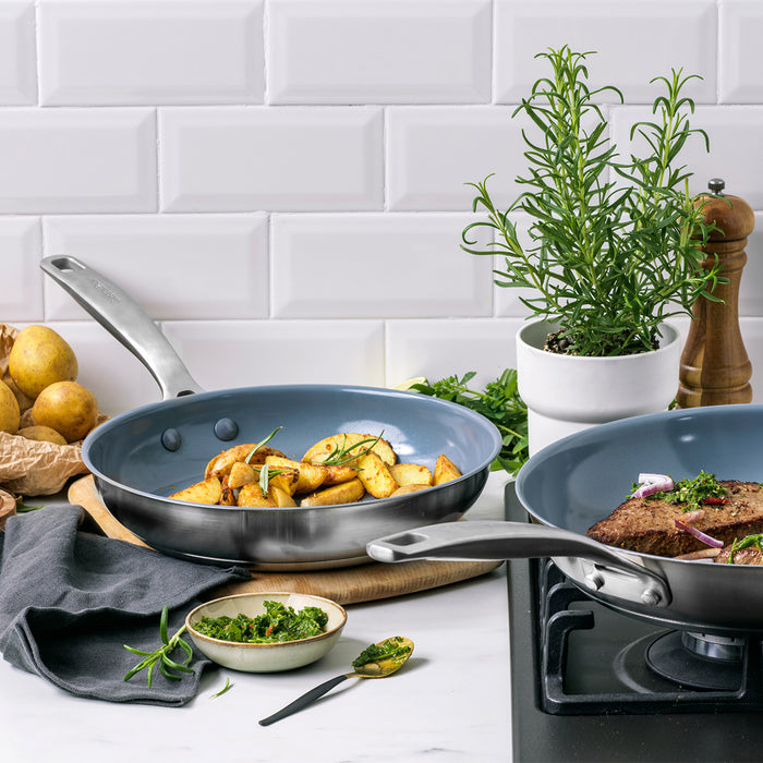 Two stainless steel nonstick pans with food, from a 10-piece cookware set, on a white countertop.