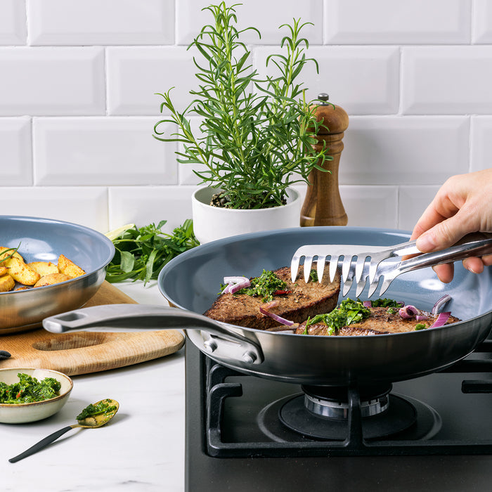Tongs holding patties with greens and onions in a stainless steel nonstick frypan on a stovetop.