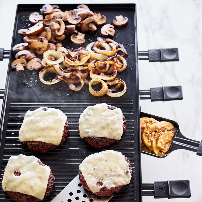 Four cheeseburgers topped with onions, mushrooms, cheese, and pineapple grilling on a countertop grill.