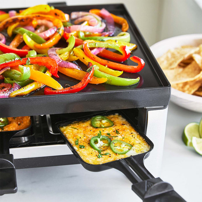 Assorted peppers grilling beside cheese, jalapeños, and tortilla chips on a flat metal grill surface.