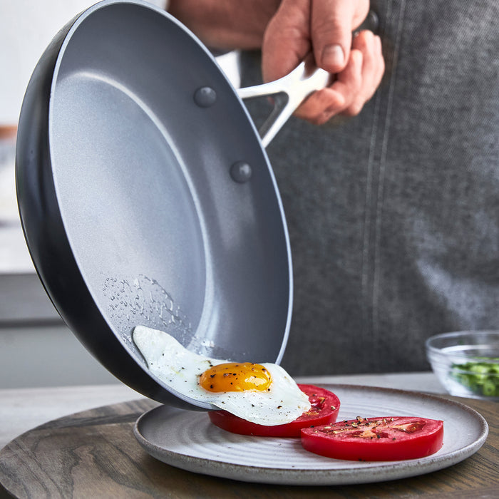 Fried egg sliding from a grey ceramic nonstick frying pan with lid onto sliced tomatoes on a plate.
