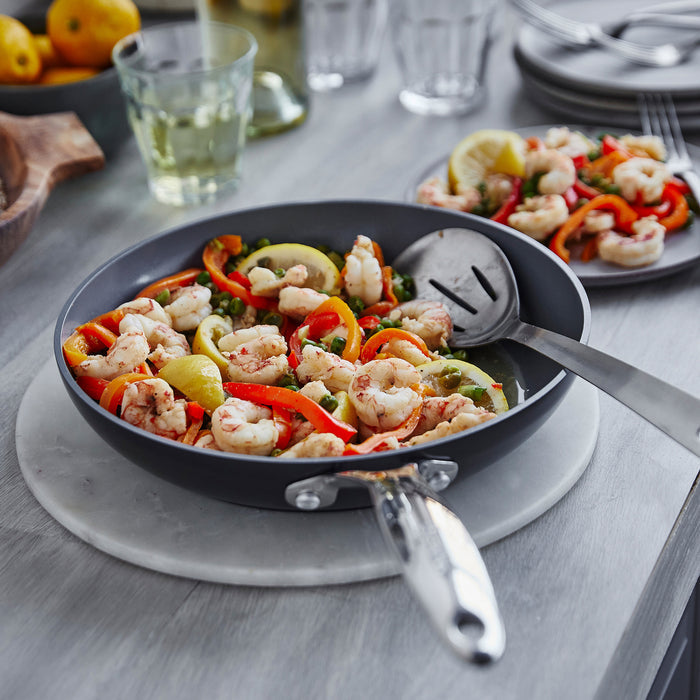 Shrimp, bell peppers, and lemon in a gray ceramic nonstick frypan with glass lid on a tabletop.