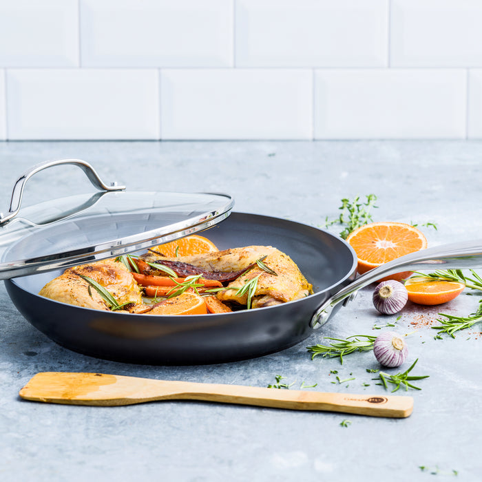 Gray ceramic nonstick frypan with lid, holding chicken, carrots, and herbs; garlic, orange slices, and spatula nearby.