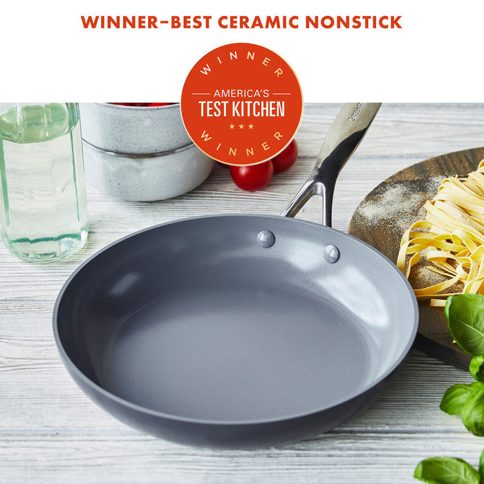 Gray ceramic nonstick frypan on wood surface next to pasta, tomatoes, and stacked white bowls.