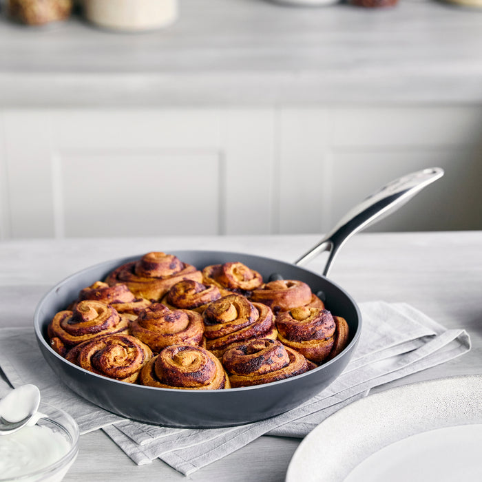 Cinnamon rolls in a pan on a gray cloth beside a spoon, plate, and Valencia Pro ceramic nonstick frypan.
