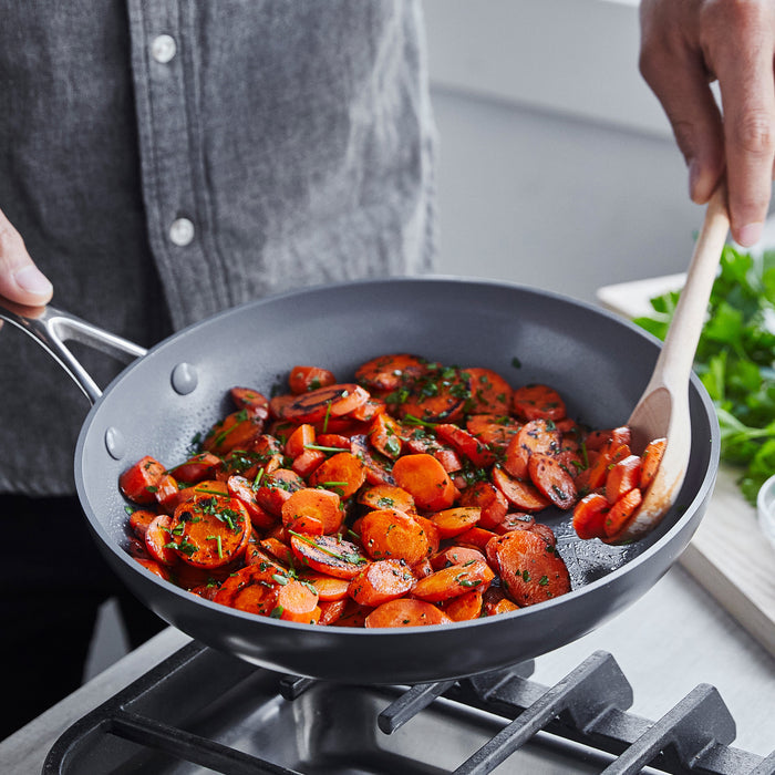Sliced carrots and herbs being stirred with a wooden spoon in a 10-inch ceramic nonstick frypan.