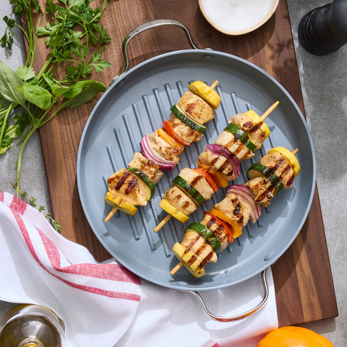 Grilled chicken and veggie kebabs on a round ceramic nonstick grill pan, with fresh herbs next to the pan.