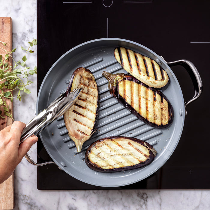Tongs turning eggplant on a round ceramic nonstick grill pan with assorted vegetables.