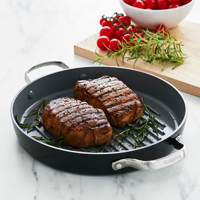 Two grilled steaks with rosemary in a gray ceramic grill pan, cherry tomatoes and herbs on the side.