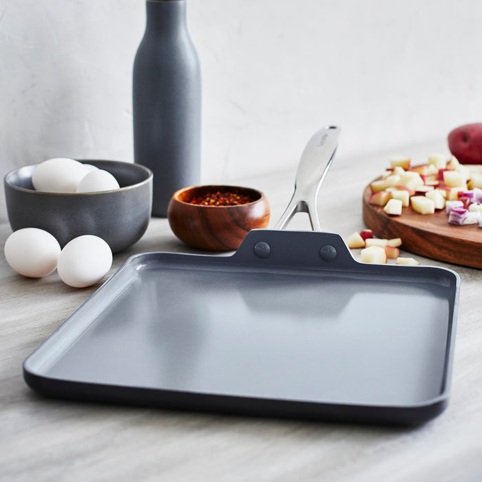 Square ceramic nonstick griddle on a counter, with eggs, spices, and chopped vegetables placed nearby.
