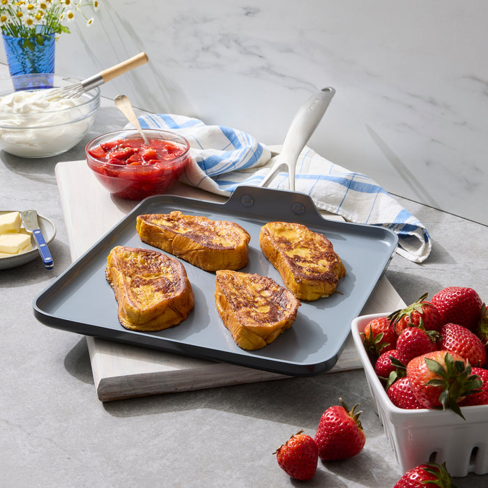 Four slices of French toast on a square ceramic nonstick griddle, with strawberries and yogurt beside the pan.