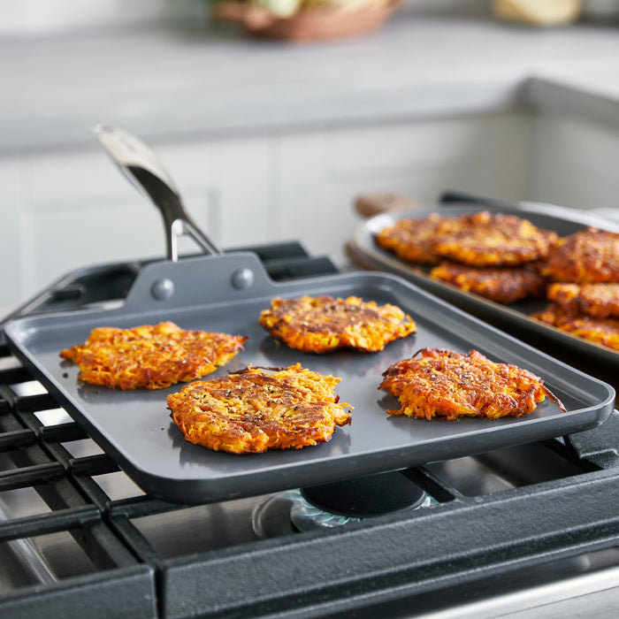 Three potato pancakes cooking on a gray ceramic nonstick square griddle, with plated food in the background.