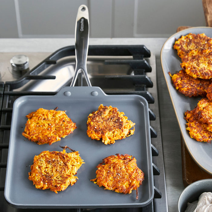 Four vegetable fritters cooking on a square ceramic nonstick griddle, with other breakfast foods visible nearby.