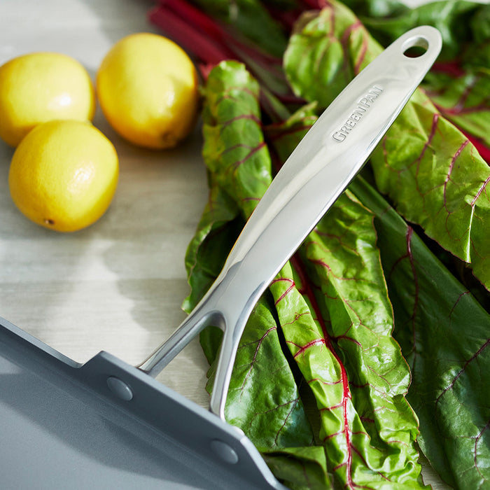 Square ceramic nonstick griddle on leafy greens next to three lemons on a light surface.
