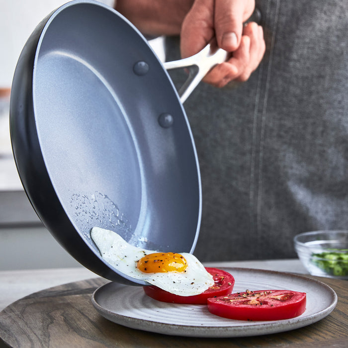 Hand sliding a sunny-side-up egg from a ceramic nonstick frypan onto sliced tomatoes on a plate.
