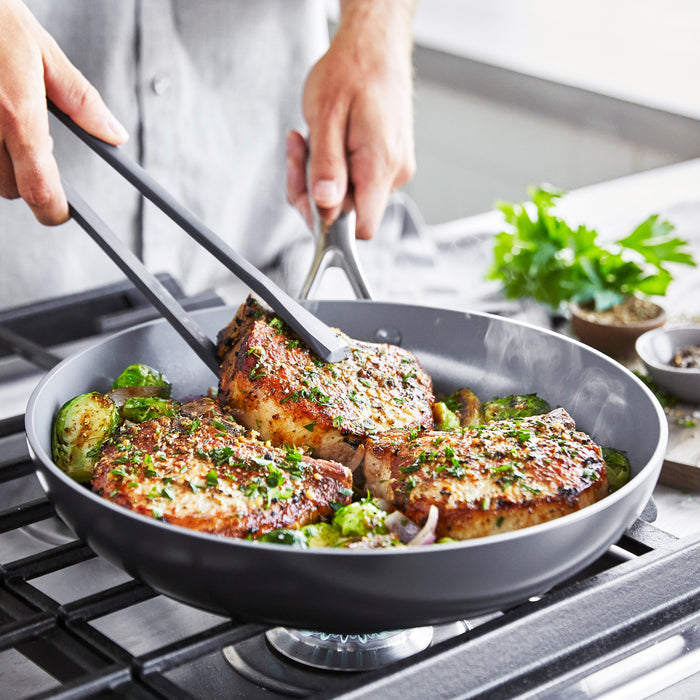 Seasoned pork chops and vegetables cooking in a grey Valencia Pro ceramic skillet on a stovetop, held with tongs.